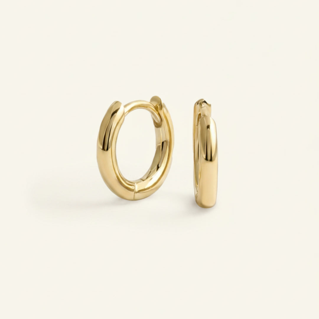 Pair of bold 18K gold hoop earrings standing upright on a soft off-white #F0EDE7 background, polished with a high-gloss finish, photographed in luxury jewelry style.