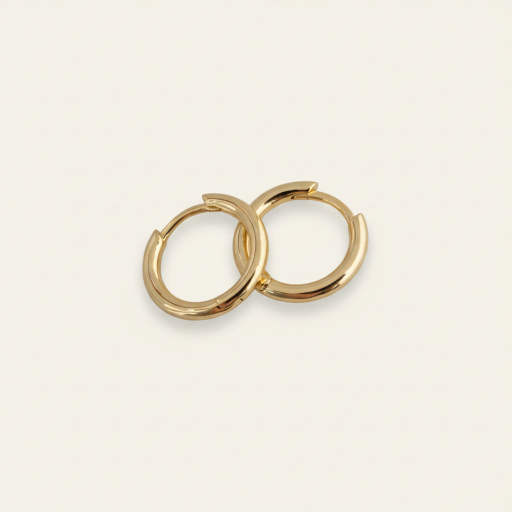 Pair of minimalist 18K gold hoop earrings displayed on a soft off-white #F0EDE7 background, polished to a mirror finish, photographed in luxury jewelry style.