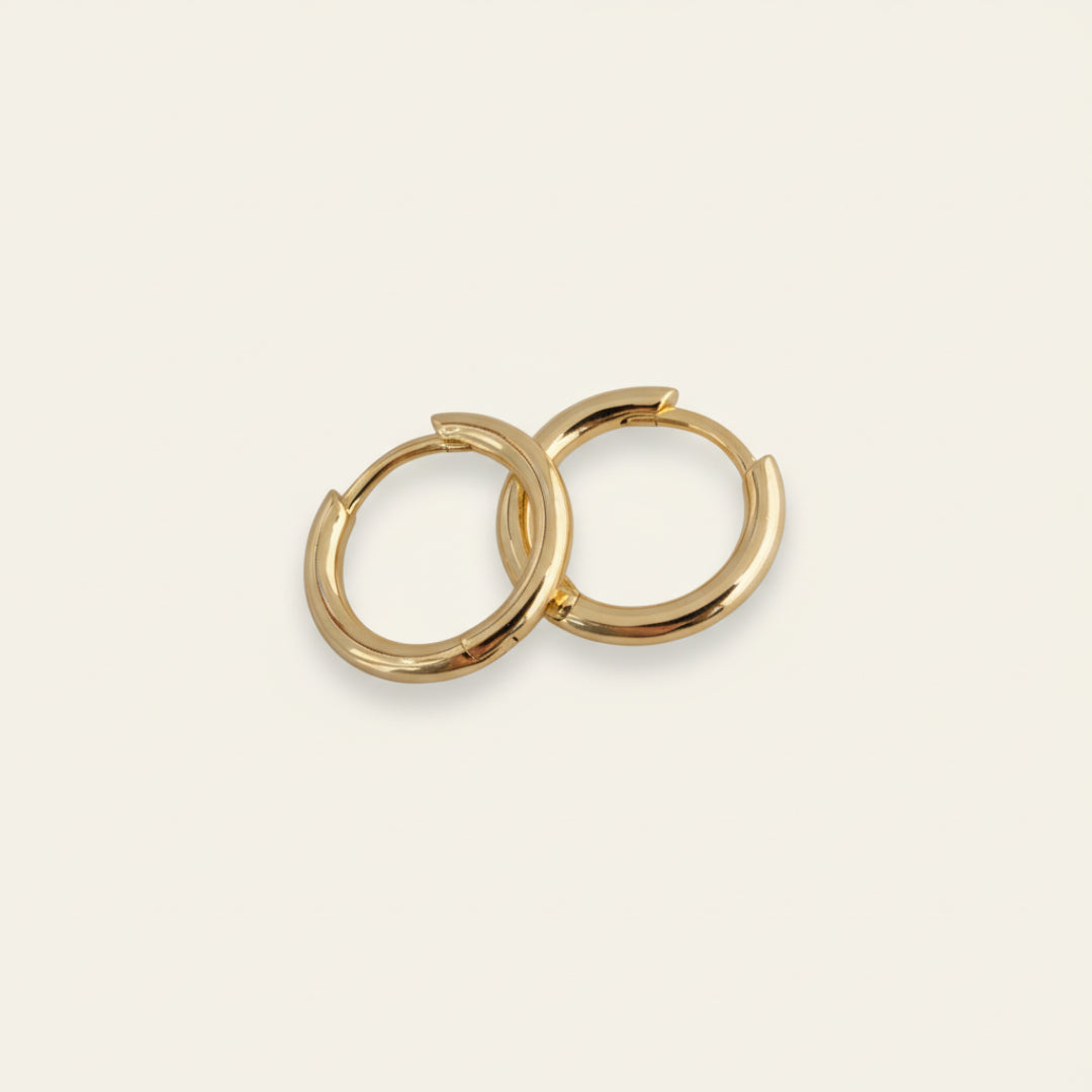 Pair of minimalist 18K gold hoop earrings displayed on a soft off-white #F0EDE7 background, polished to a mirror finish, photographed in luxury jewelry style.