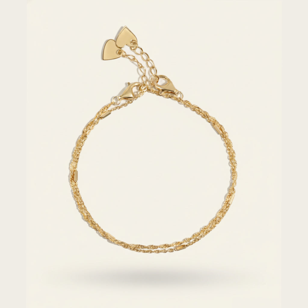 Delicate 18K gold rope chain bracelet featuring dual heart clasps and adjustable links, displayed on a soft off-white #F0EDE7 background, photographed in luxury jewelry style.