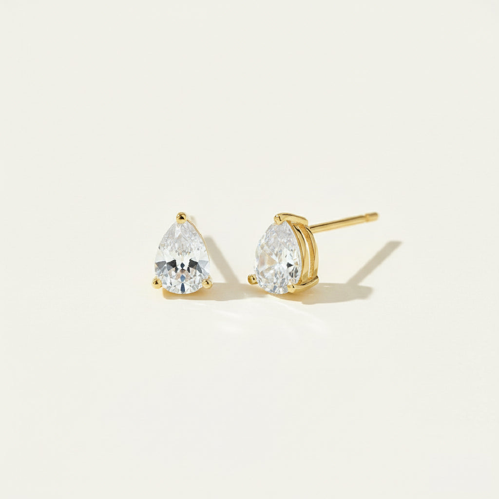 Pair of gold teardrop diamond stud earrings shown in side and front view, featuring brilliant-cut gemstones in a minimalist prong setting, displayed on a soft off-white #F0EDE7 background in luxury jewelry style.