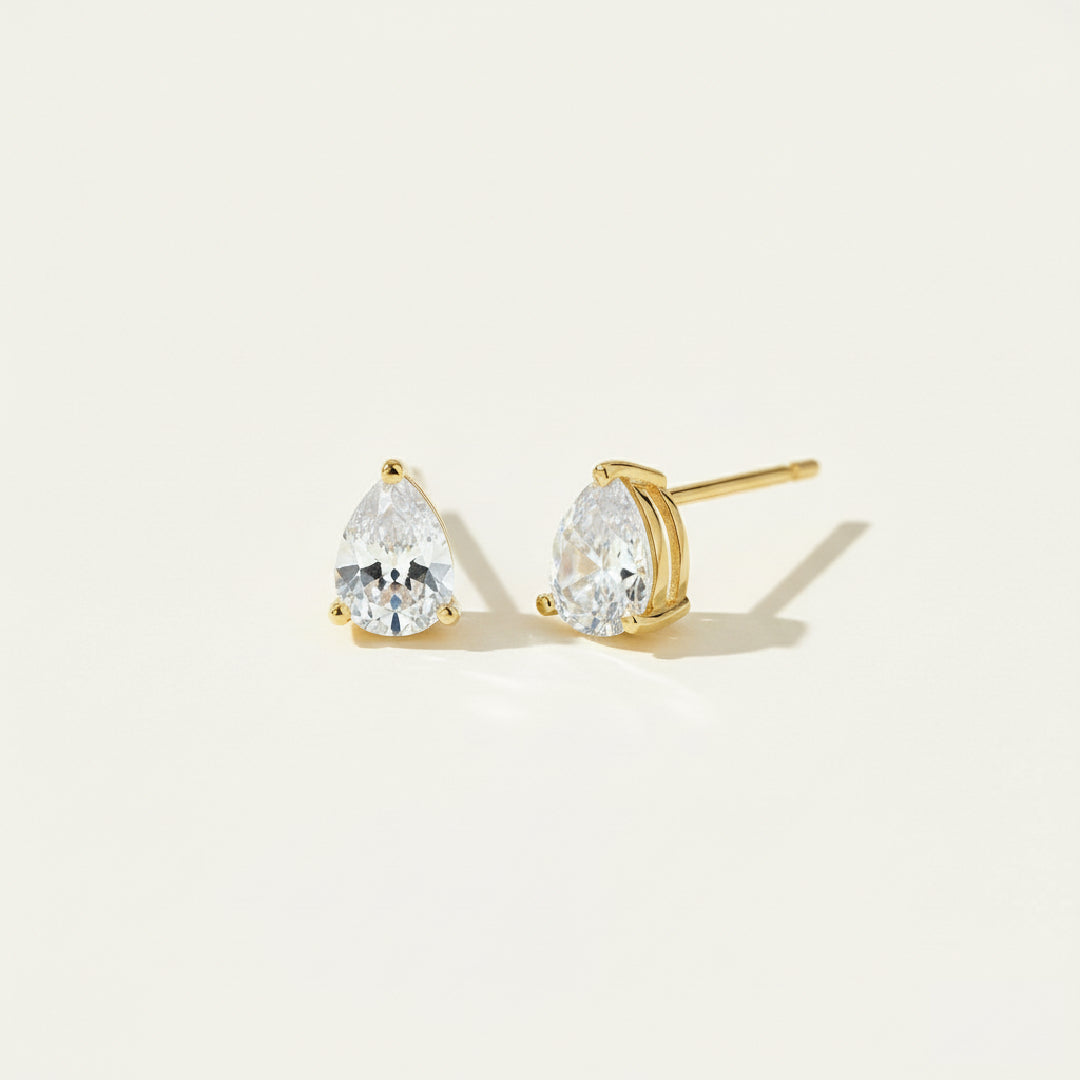 Pair of gold teardrop diamond stud earrings shown in side and front view, featuring brilliant-cut gemstones in a minimalist prong setting, displayed on a soft off-white #F0EDE7 background in luxury jewelry style.
