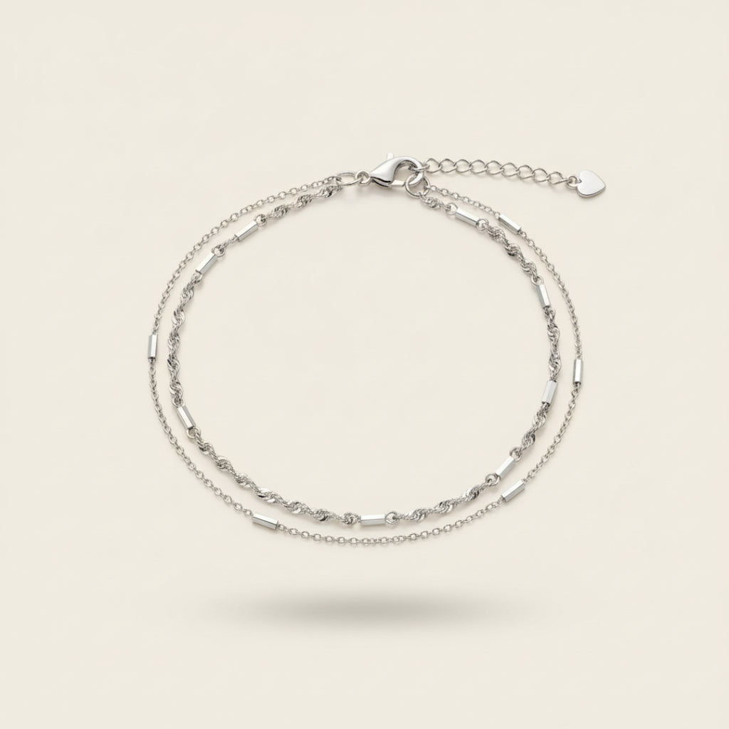 Top view of the Minimalist Double Bracelet Set in sterling silver featuring delicate dual chains with polished bar details and heart clasp, displayed on a soft off-white #F0EDE7 background in luxury jewelry photography style.