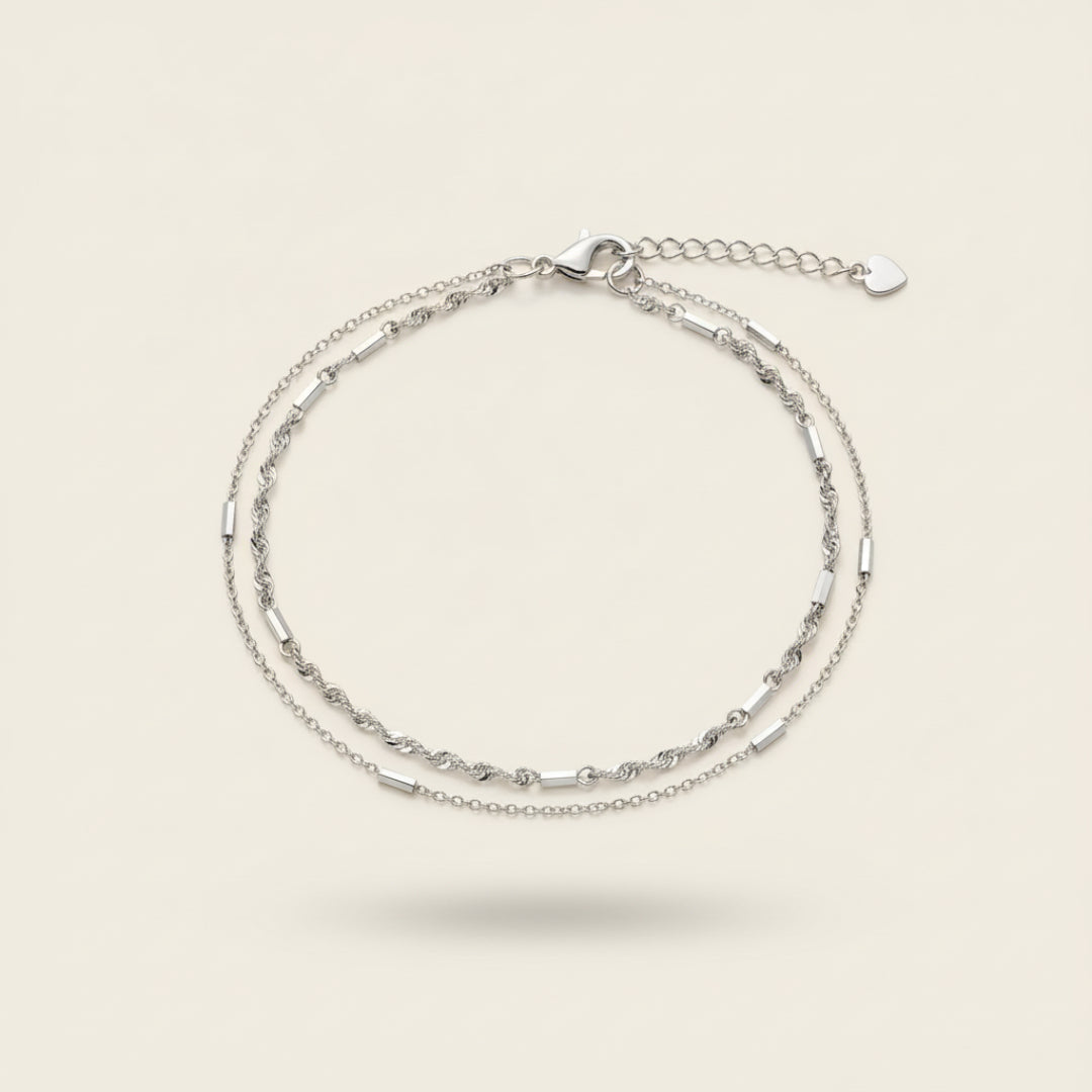 Top view of the Minimalist Double Bracelet Set in sterling silver featuring delicate dual chains with polished bar details and heart clasp, displayed on a soft off-white #F0EDE7 background in luxury jewelry photography style.