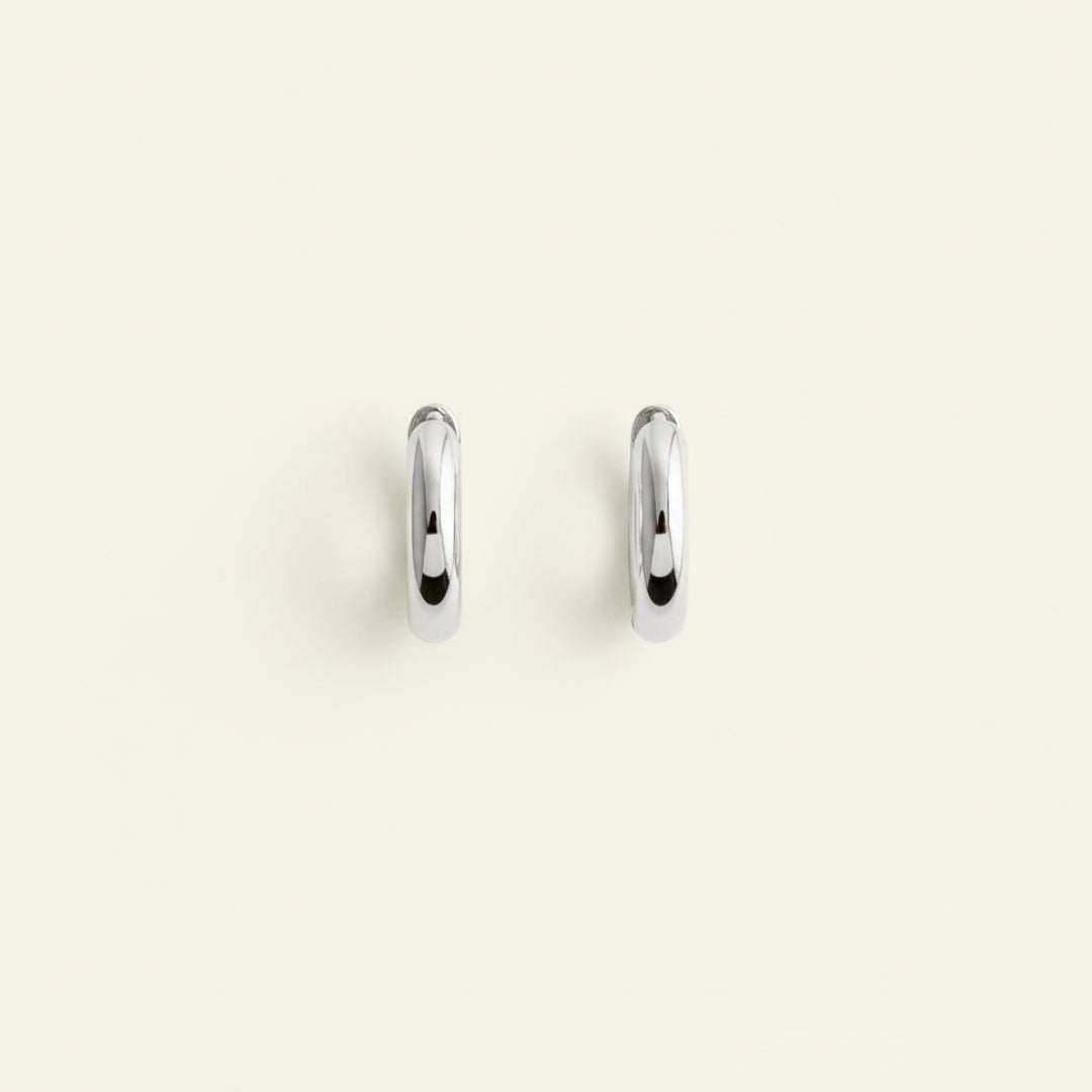 Pair of polished sterling silver hoop earrings displayed upright on a soft off-white #F0EDE7 background, featuring a smooth reflective finish, photographed in luxury jewelry style.