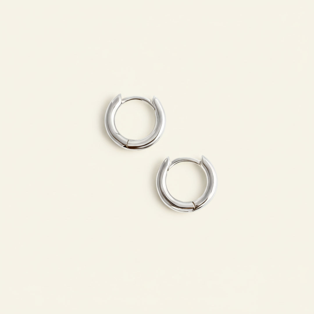 Pair of minimalist sterling silver hoop earrings displayed on a soft off-white #F0EDE7 background, polished with a mirror finish, captured in high-quality luxury jewelry style.